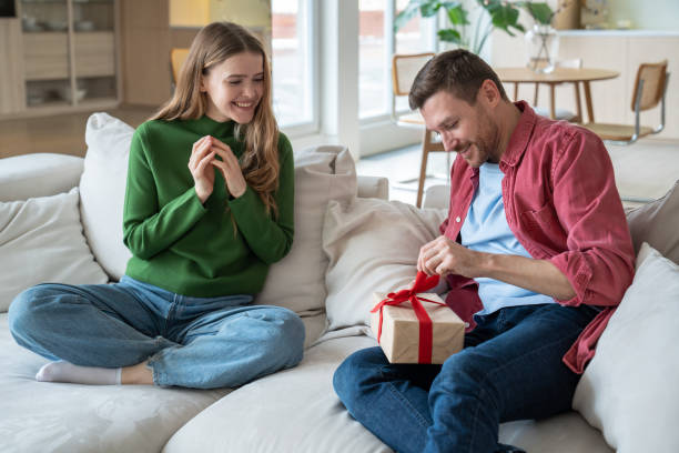 Joyful birthday celebrator sitting next to glad woman, unbracing ribbon, going to unwrap giftbox Thrilled intrigued man unbracing red ribbon, trying to unwrap cardboard giftbox. Pleasantly smiling merry loving woman looking with excitement and hope that beloved darling man will like present gift for girlfriend stock pictures, royalty-free photos & images