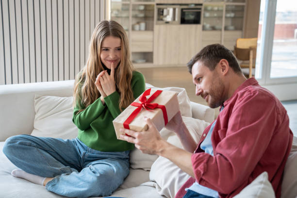 Intrigued man sitting on sofa, trying to unwrap giftbox, and glad woman looking at him with love Pleasantly smiling merry European woman looking with excitement as intrigued interested man trying to unwrap giftbox with red ribbon. Unexpected surprise for birthday, anniversary, New Year unusual couple stock pictures, royalty-free photos & images