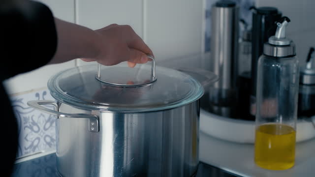Water Boils In Large Iron Pot On The Stove while woman opens up the Lid to release steam in slow motion. Close up of Bubbles Of Boiling Water in the kitchen during a day. Sony FX3 in 4K.
