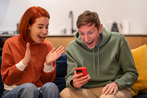 Excited overjoyed millennial happy young family couple looking at phone screen screaming with joy while sitting on sofa, happy man and woman holding smartphone celebrating triumph, winning lottery