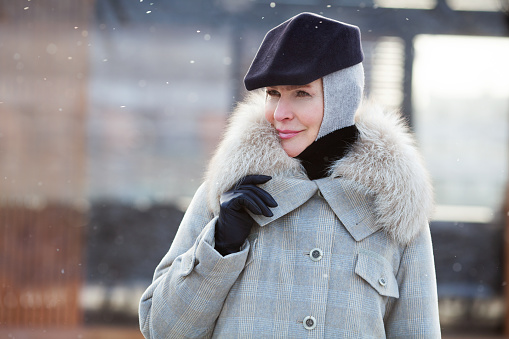 Portrait trendy mature woman Stands outside in snow, wears fashionable winter multilayer outfit, grey coat with fur collar, hood and stylish black cap. Female warm clothes.