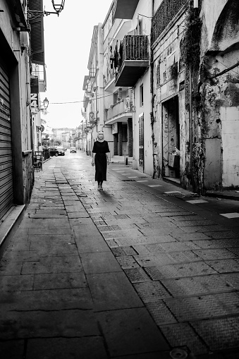 Monochrome image of a mature woman in a black dress walking along the alleys of the town of Scafati, south of Naples, Italy.