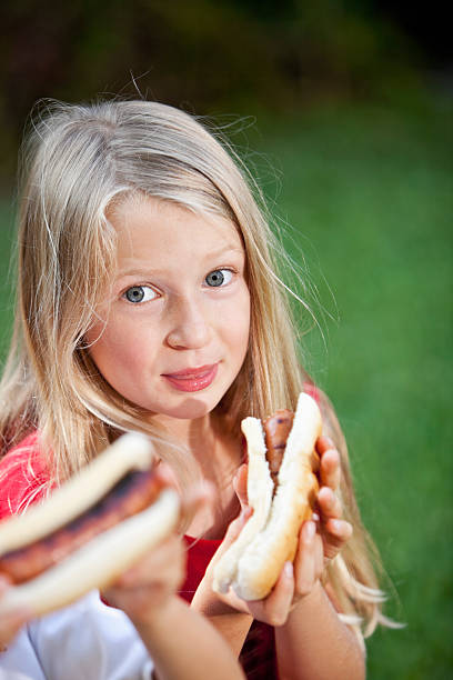 Young cute european girl in t-shirt is eating unhealthy food like hot dog and chips at sunny day