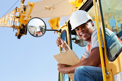 Low angle view of a young African American man sitting in the cab of a crane, looking through the door, down at the camera, smiling.  He is wearing a white hard hat, orange safety vest and jeans.  He has a clipboard in one hand and is hanging on with the other.