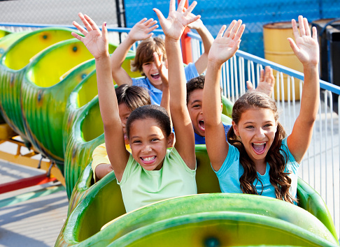 A group of five multiracial children riding a green roller coaster.  They are having fun, arms raised up in the air.  The focus is on the two girls in front, 11 years old.