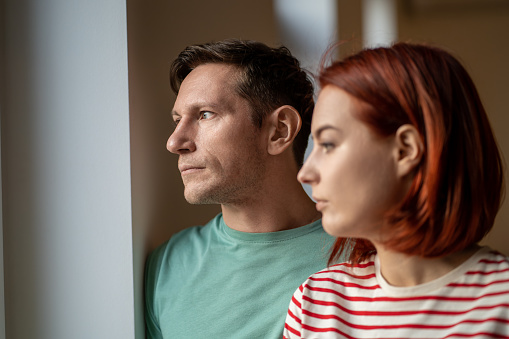 Pensive middle aged german man and woman looking out the window thinking about relationship troubles. Closeup of sad caucasian couple looking anxiously into future, each thinking about life problems