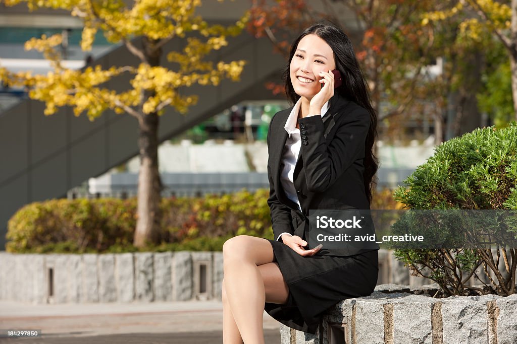 Asian Female in Suit Uses Cell Phone Horizontal shot of attractive young adult asian woman in business attire sits on a stone planter in a business plaza in Tokyo.  Her legs are crossed and she holds her cell phone to her ear and smiles. 20-24 Years Stock Photo
