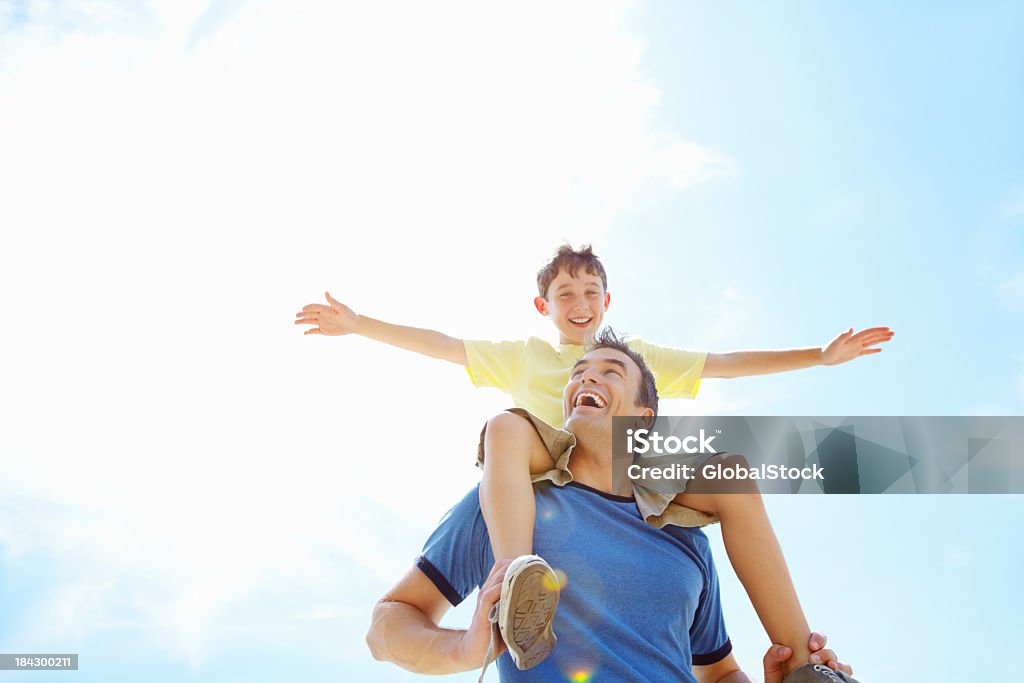 Son having fun sitting on his father's shoulders Low angle view of cheerful father carrying his son on shoulder against cloudy sky Carrying On Shoulders Stock Photo