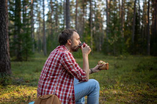 Man hiker drinking tea from mug during hiking in forest. Male during small halt eating sandwich and drinking coffee in autumn woodland, resting stopped for break after walk in woods.