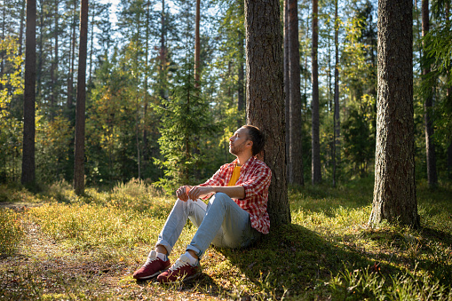 Man camper tourist resting sitting near tree in forest with closed eyes in warm sunny day. Guy enjoying in unity with nature in park. Male relaxing dreaming meditating, tourism travel hiking concept.