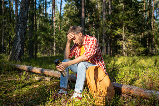 Tired man hiker having break in mountains feeling bad from height sitting on log in forest with thermos in hands. Hiking camping, dizziness, lack of air, health problems, breathing difficulty concept.