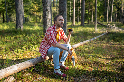 Man camper hiker sitting on log in forest enjoying drinking tea from thermos having break. Tourist guy on halt in summer day. Camping, hiking tourism concept. Outdoors activity recreation in woodland.