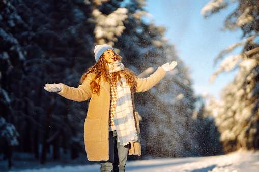 Young woman in warm clothes plays with snow on a sunny winter day. Happy female tourist enjoying a snowy day in the park. Concept of walking, fun.