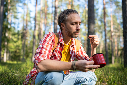 Man eating berries in nature. Thoughtful unsmiling middle aged male bent over mug in hand. Guy found wild natural gifts berries and herbs in order to enjoy freshly brewed black tea. Stress relieve