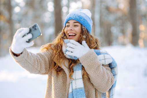 Happy woman in warm clothes with a phone in her hands having fun on a winter snowy day. A young woman takes a selfie on her phone in a snowy park. Fun concept. Active lifestyle.