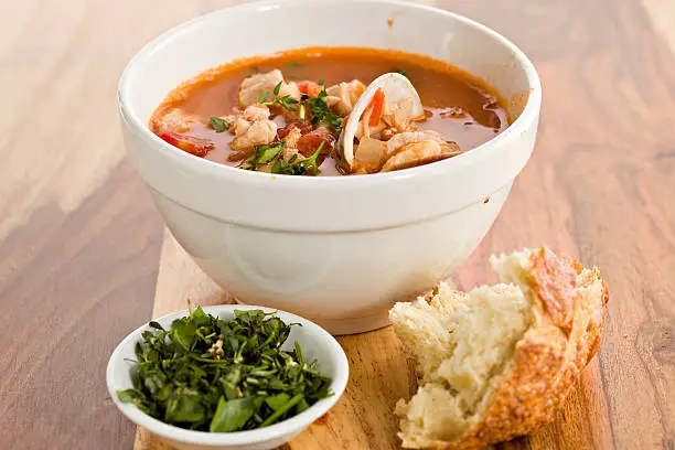 "A close up of a white bowl full of red seafood soup and in the foreground we have a chunk of crusty San Francisco Style sourdough bread and a small white bowl containing chopped parsley,basil and thyme. Shot against  a wooden surface."