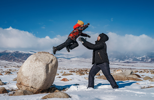 Child jumping on daddy from big wall on mountains background at winter season
