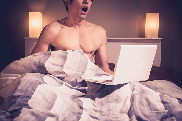 Young man in bed watching pornography on laptop stock photo