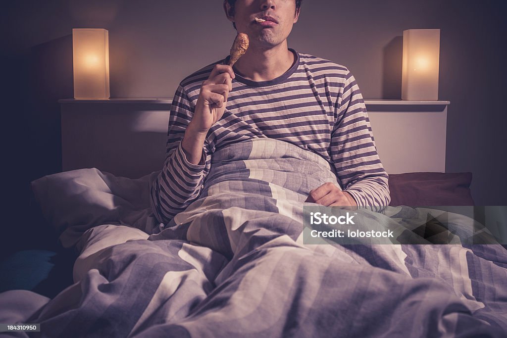 Young man is sitting in bed and eating chicken Young man is sitting in bed at night and eating a chicken drumstick Bed - Furniture Stock Photo