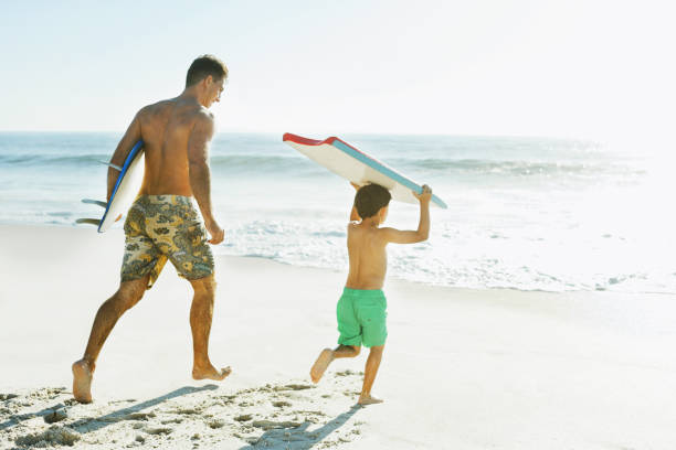 padre e figlio per il trasporto di tavola da surf e il bodyboard sulla spiaggia - men beach running shirtless foto e immagini stock