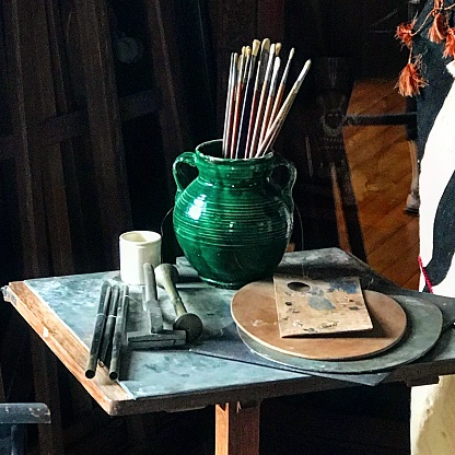 This photograph shows a collection of art supplies, including brushes in a green ceramic jug, a palette, and various drawing tools, arranged on a table with a marble-like surface.