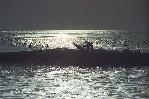 This photograph captures a silhouetted scene of a surfer shreding a wave in Malibu, with another performing a maneuver on a wave, against the backdrop of a shimmering sea surface under what appears to be a setting or rising sun.