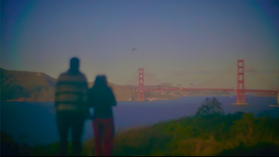 This photograph depicts a blurred image of two people looking towards the Golden Gate Bridge in the distance.