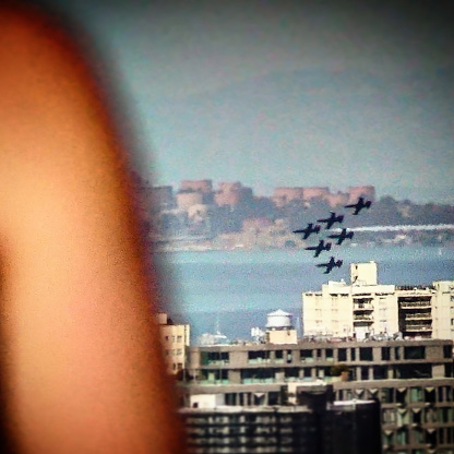 This photograph captures a blurred view of a city skyline with a formation of US Navy aircraft flying over the water during a Fleet Day event in San Francisco.