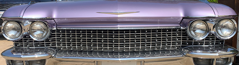 This photograph shows the front grille and headlights of a classic Cadillac, featuring its distinctive chrome details and emblem, with a California license plate visible below.