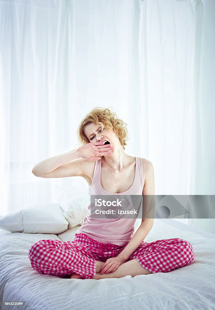 Awakening Young adult woman wearing pajamas sitting cross-legged on the bed in a bedroom and yawning.  Women Stock Photo