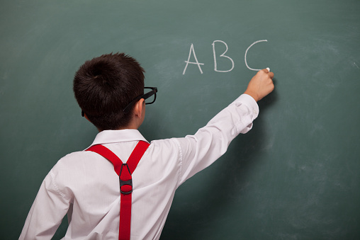 Back view of little boy wearing white shirt and red suspenders writing alphabet on green blackboard