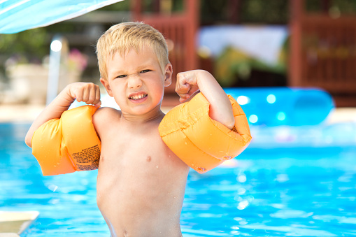 Cute little boy with water wings standing by the swimming pool and showing his muscles and his strenght.