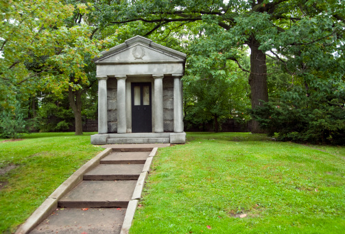 A mausoleum in an urban setting surrounded by green grass and large deciduous trees in the summer months.