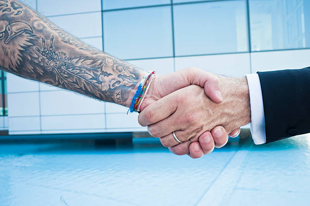 Arm with tattoos and arm in suit shaking hands Close-up of a handshake between a businessman and a tattooed man showing just the hands and arms. There is a skyscraper in the background. arm tattoo ideas for men pictures stock pictures, royalty-free photos & images