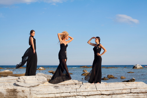Three beautiful ladies in the dresses standing on the rocks on the sea lagoon