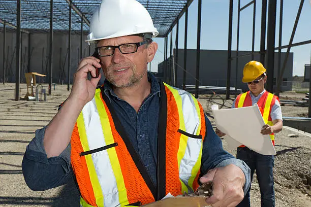 A safety inspector making a phonecall while an engineer behind him reviews plans.