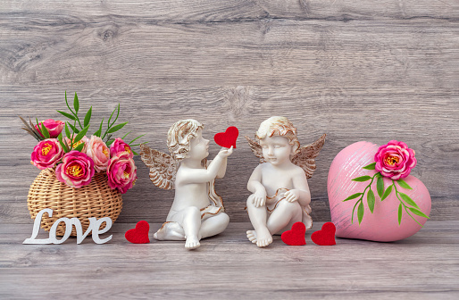 Valentine's day. The angel statue is decorated with a decorative heart and flowers in a basket. The letters of the word Love. A gift for lovers. Beige wooden background.