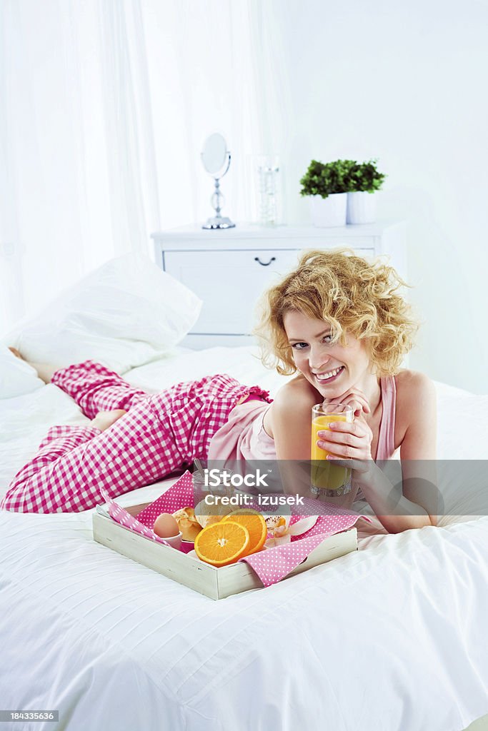 Healthy breakfast in bed Happy young adult woman lying on a bed and going to eat breakfast. Holding a glass of orange juice, looking at camera and smiling. 25-29 Years Stock Photo