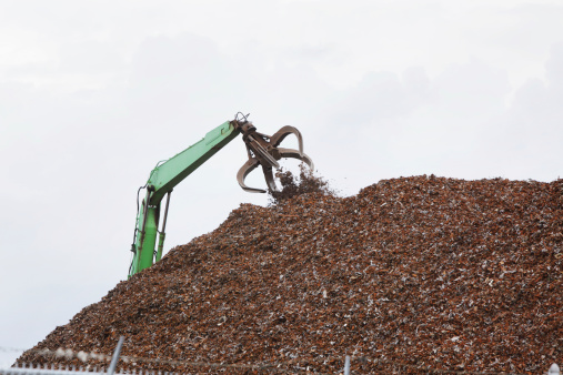Tall crane with a hydraulic-operated claw attachment dumps additional material onto a mountain of shredded scrap metal behind a barbed wire chainlink fence at a Portsmouth, New Hampshire recycling center.
