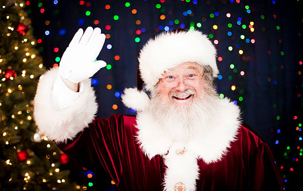 Santa Claus Waving Portrait of a real Santa Claus waving and smiling background with lots of Christmas lights and a decorated Christmas tree. Room for copy space. Merry Christmas! kruis stock pictures, royalty-free photos & images
