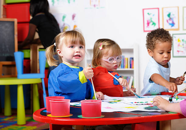 Group Of Toddlers Painting While Their Carer Tidies A portrait of a group of  toddlers painting whilt their carer tidies. two groups stock pictures, royalty-free photos & images