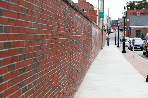 Diminishing perspective view of a long brick wall and sidewalk on an older New England city street in Portsmouth, New Hampshire. An unrecognizable person strolls along the sidewalk in the far distance.