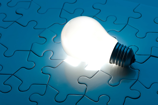An illuminated light bulb is sitting on top of a plain, dark blue colored puzzle, consisting of about two dozen puzzle pieces.  The light bulb is lying on its side, resting where there is a missing puzzle piece.  It is not screwed in to any source.