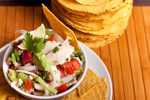 Photograph with warm tones and taken from a high angle of a bowl full of freshly made ceviche garnished with a wedge of avocado and cilantro; sitting on bamboo place mat with copy space left at right side of frame 
