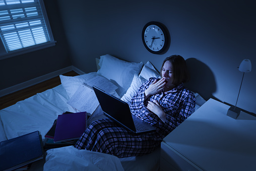 A young woman college or university student with insomnia or a deadline, working and studying late into the night with her laptop computer and homework in her dorm room bed. She wears plaid pajamas and yawns with fatigue as she faces the anxiety and frustration, stress and pressure of exams.