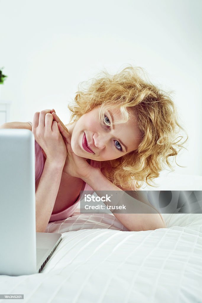 Using computer in a bed Smiling young adult woman lying on stomach on a bed and using laptop, looking at camera. 25-29 Years Stock Photo