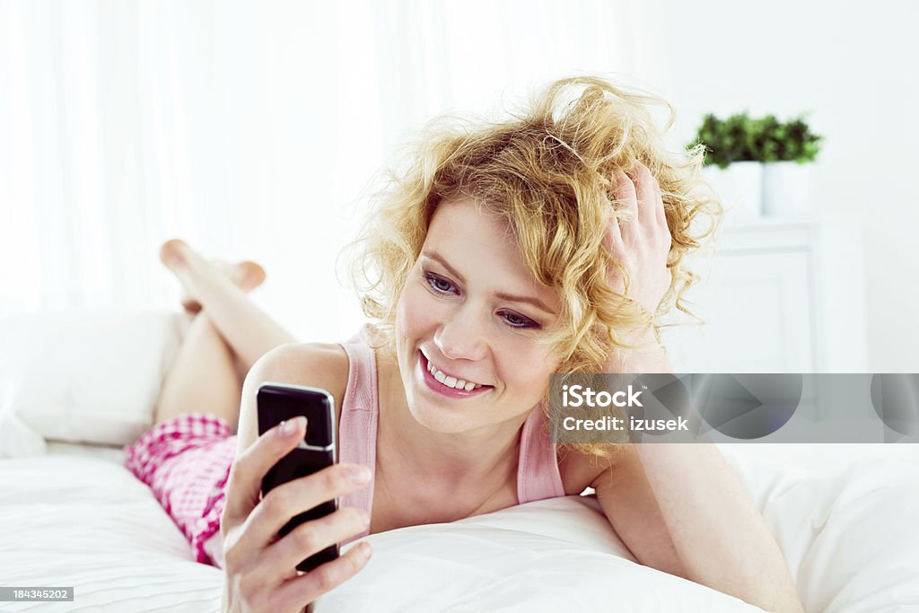 Using cell phone in a bed Smiling young adult woman lying on stomach on a bed and texting on her mobile phone. 20-24 Years Stock Photo