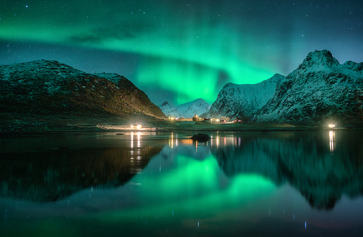 Aurora borealis, snowy mountains, sea, fjord, reflection in water, street lights at starry winter night. Lofoten, Norway. Northern lights. Landscape with polar lights, snowy rocks, sky with stars