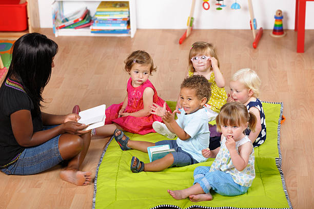 childminder чтение чтобы группа маленьких детей в детском саду - baby1 стоковые фото и изображения