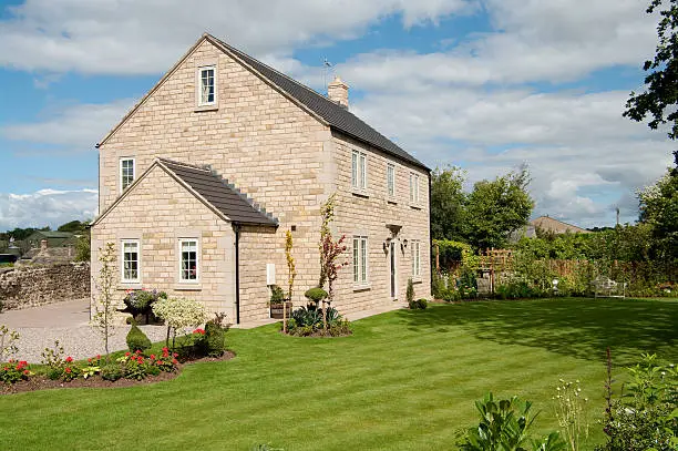 Attractive newly built English three-storey cottage built in stone in a traditional style. Striped lawn with topiary forms in newly established garden. Summer day. The house is detached and is set in its own grounds.